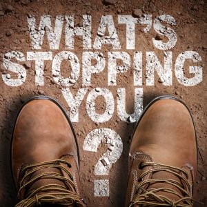 Top-down view of worn brown boots standing still on a dirt path with the phrase 'What’s Stopping You?' painted on the ground